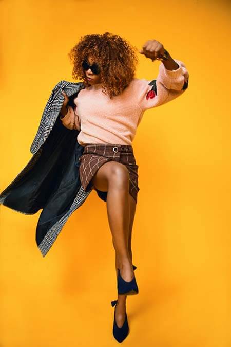 Black womxn with a professional afro hairstyle standing in front of an orange-yellow background with one foot and arm lifted in the air.