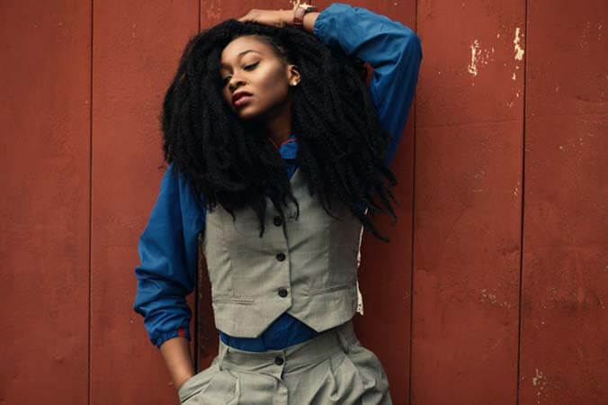 Black womxn with a professional havana twists afro hairstyle wearing blue shirt, grey suit set, in front of a red background